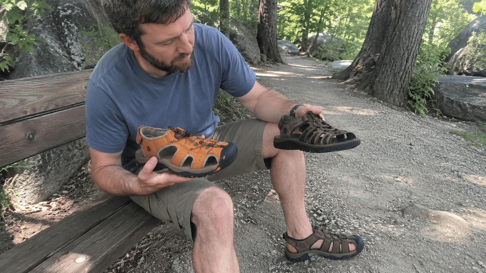 A man comparing two all-terrain sandals side by side on a wooded trail